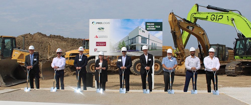 9 individuals standing in front of heavy machinery holding shovels at a groundbreaking event